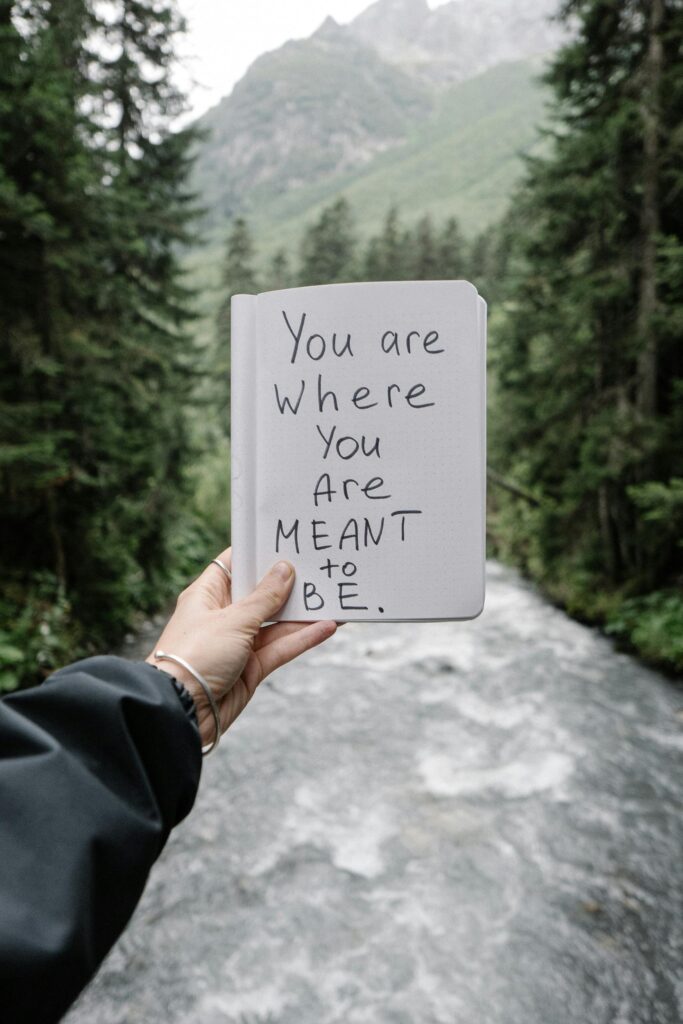 Hand holding a motivational quote by a forest river, evoking mindful travel and introspection.